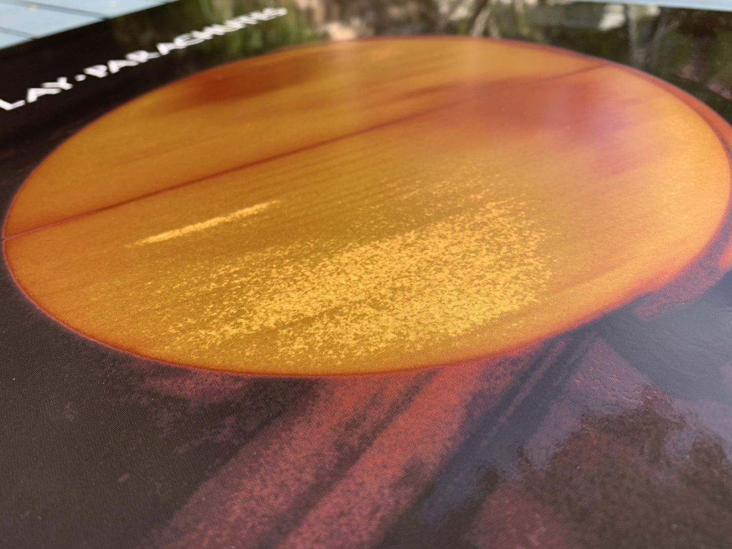 Parachutes Lithograph in a cozy living room, hung above a wooden, vintage, caramel brown leather sofa. To its left, there’s a tall leafy plant in a pot, adding a touch of nature. The wall is beige. The poster is on a black frame. The artwork’s yellow, ethereal glow complements the sofa’s earthy tones